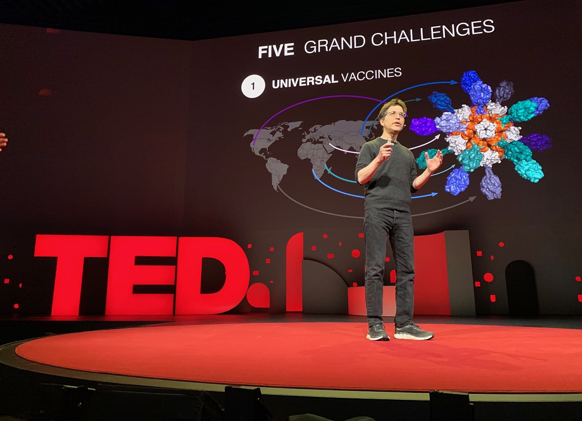 David Baker, director of the Institute for Protein Design, speaks on the TED2019 stage in front of a slide reading Five Grand Challenges, Universal Vaccines, with a rendering of a designed protein nanoparticle, April 2019. Illustrative image, not from the RFD4-Proteina announcement. Photo: Steve Jurvetson, CC BY 2.0, via Wikimedia Commons. Source page: https://commons.wikimedia.org/wiki/File:Audacious_Philanthropy_Congrats_to_David_Baker_and_his_Institute_for_Protein_Design.jpg