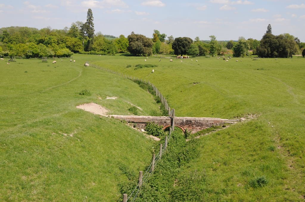 Rolling green parkland with grazing sheep, a fenced stream and a small brick bridge, framed by mature trees, at Wimpole Park, Cambridgeshire, England, May 27, 2013. Photo: Philip Halling, CC BY-SA 2.0, via Wikimedia Commons (Geograph). Source page: https://commons.wikimedia.org/wiki/File:Wimpole_Park_-_geograph.org.uk_-_3492584.jpg