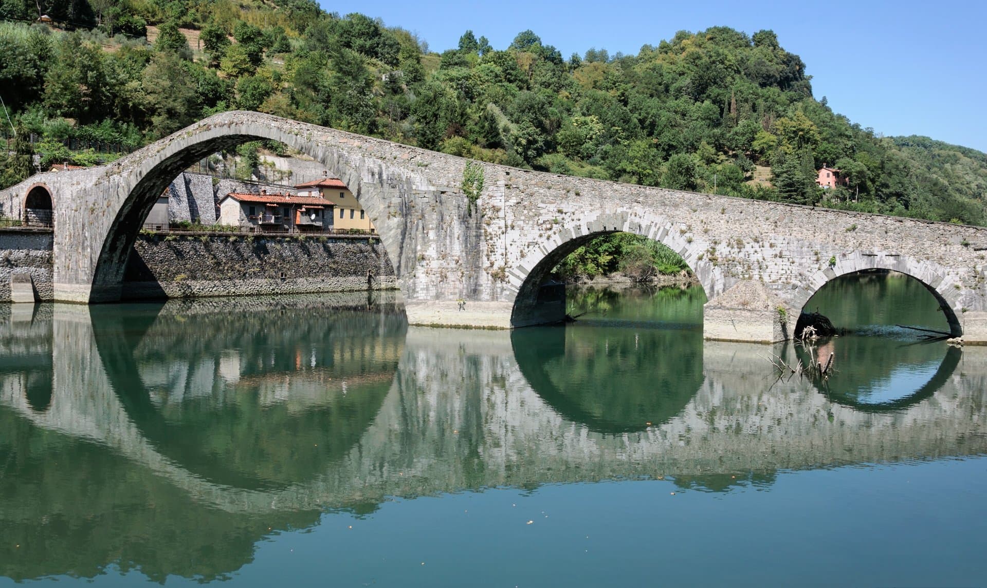The medieval Ponte della Maddalena, a stone bridge with one tall hump-backed arch and several smaller arches, reflected in still green water below wooded hills at Borgo a Mozzano, Tuscany, Aug. 23, 2011. Illustrative image of the three friends' hometown; the bridge crosses the Serchio, not the stretch of the Lima river where the ring was lost. Photo: Myrabella, CC BY-SA 4.0, via Wikimedia Commons. Source page: https://commons.wikimedia.org/wiki/File:Borgo_a_Mozzano_Ponte_della_Maddalena.jpg