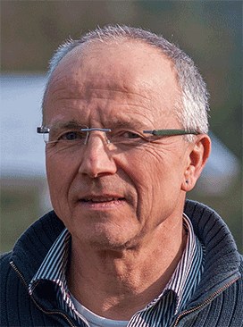 A portrait of a man with close-cropped gray hair and rimless glasses, wearing a dark zip-up jacket, outdoors.