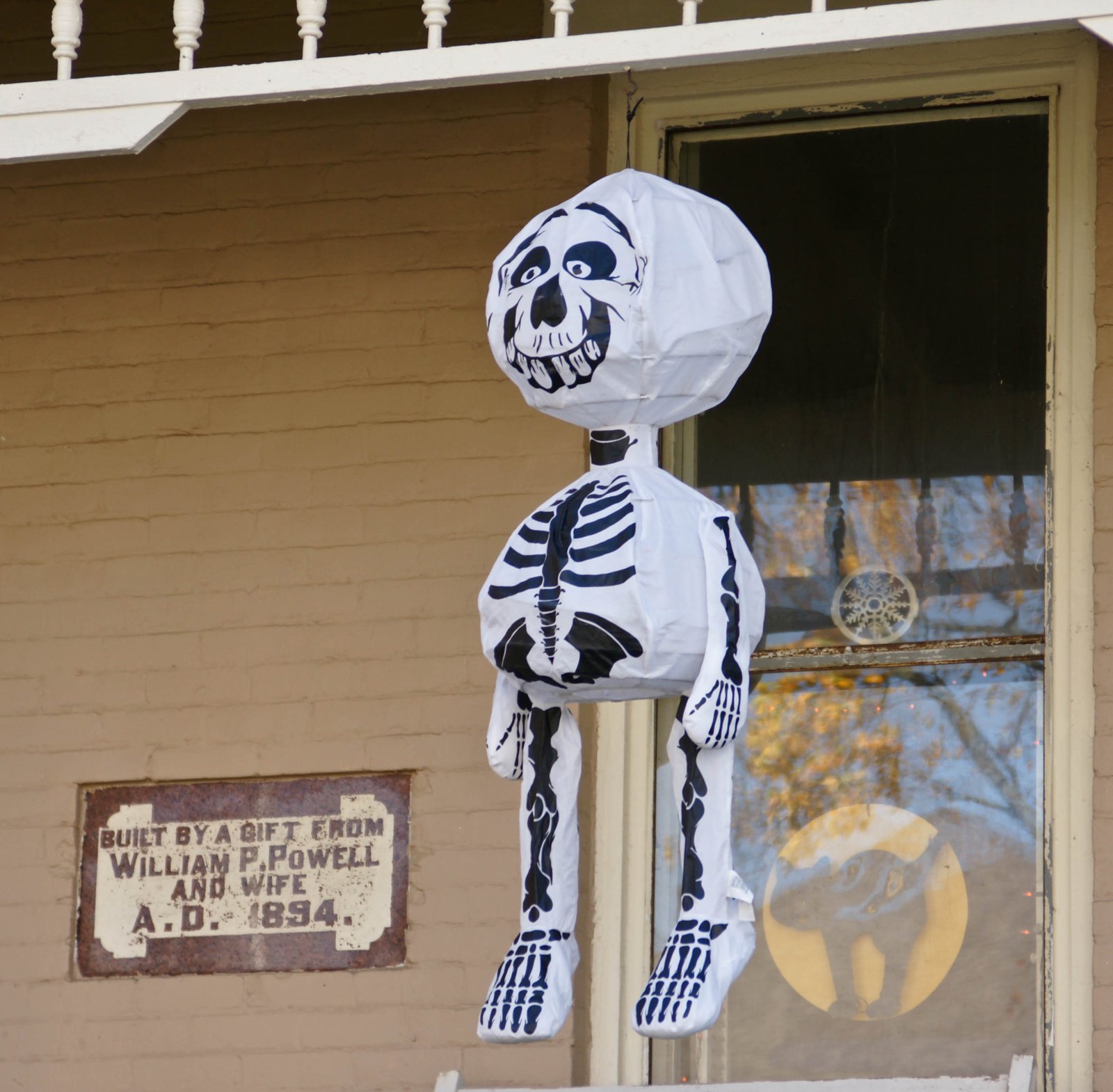 An inflatable cartoon skeleton hangs from the porch of an old brick house beside a window and a weathered plaque reading Built by a gift from William P. Powell and wife, A.D. 1894.