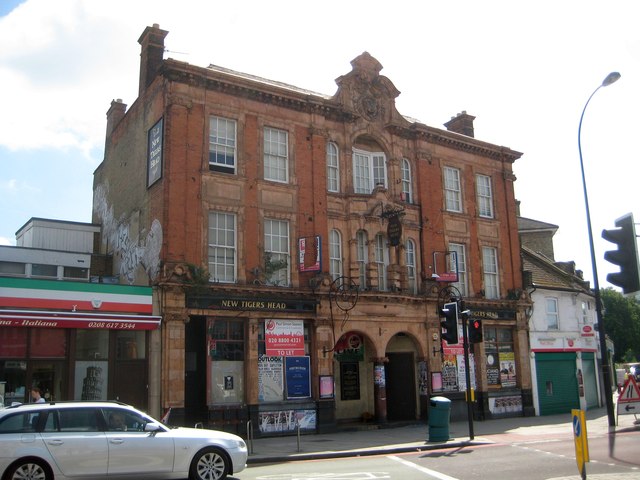 A tall red-brick Victorian building, the New Tiger's Head, on a street corner at Lee Green, with shops at street level, traffic lights and a parked silver car.