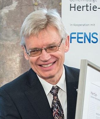 A smiling man with gray hair and glasses, in a dark suit and patterned tie, in front of an event backdrop.